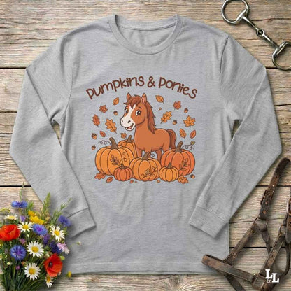 A grey long-sleeve t-shirt with a 'Pumpkins & Ponies' graphic, laid flat on a rustic wooden background with decorative autumn props including flowers, a horse bit, and a leather bridle.