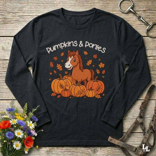 A black long-sleeve t-shirt with a 'Pumpkins & Ponies' graphic, laid flat on a rustic wooden background with decorative autumn props including flowers, a horse bit, and a leather bridle.