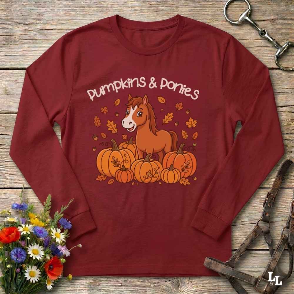A red long-sleeve t-shirt with a 'Pumpkins & Ponies' graphic, laid flat on a rustic wooden background with decorative autumn props including flowers, a horse bit, and a leather bridle.