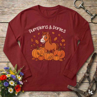 A red long-sleeve t-shirt with a 'Pumpkins & Ponies' graphic, laid flat on a rustic wooden background with decorative autumn props including flowers, a horse bit, and a leather bridle.
