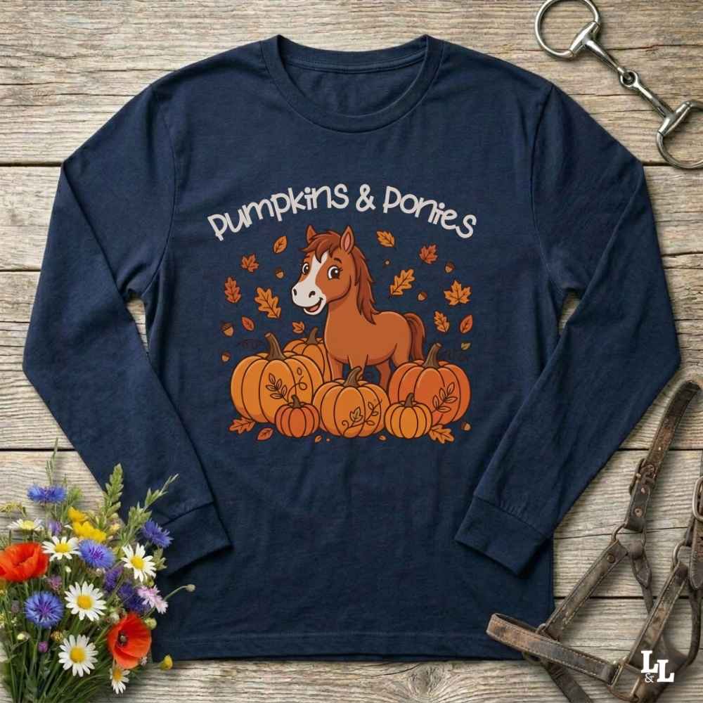 A navy blue long-sleeve t-shirt with a 'Pumpkins & Ponies' graphic, laid flat on a rustic wooden background with decorative autumn props including flowers, a horse bit, and a leather bridle.