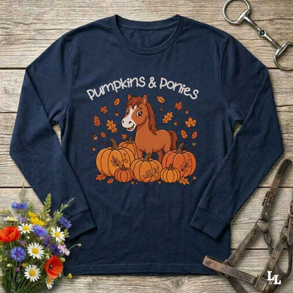 A navy blue long-sleeve t-shirt with a 'Pumpkins & Ponies' graphic, laid flat on a rustic wooden background with decorative autumn props including flowers, a horse bit, and a leather bridle.