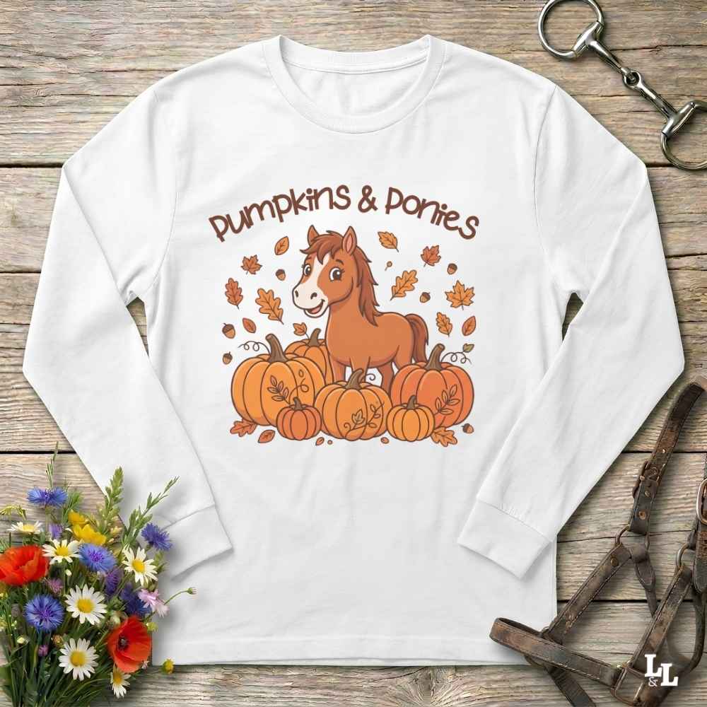 A white long-sleeve t-shirt with a 'Pumpkins & Ponies' graphic, laid flat on a rustic wooden background with decorative autumn props including flowers, a horse bit, and a leather bridle.