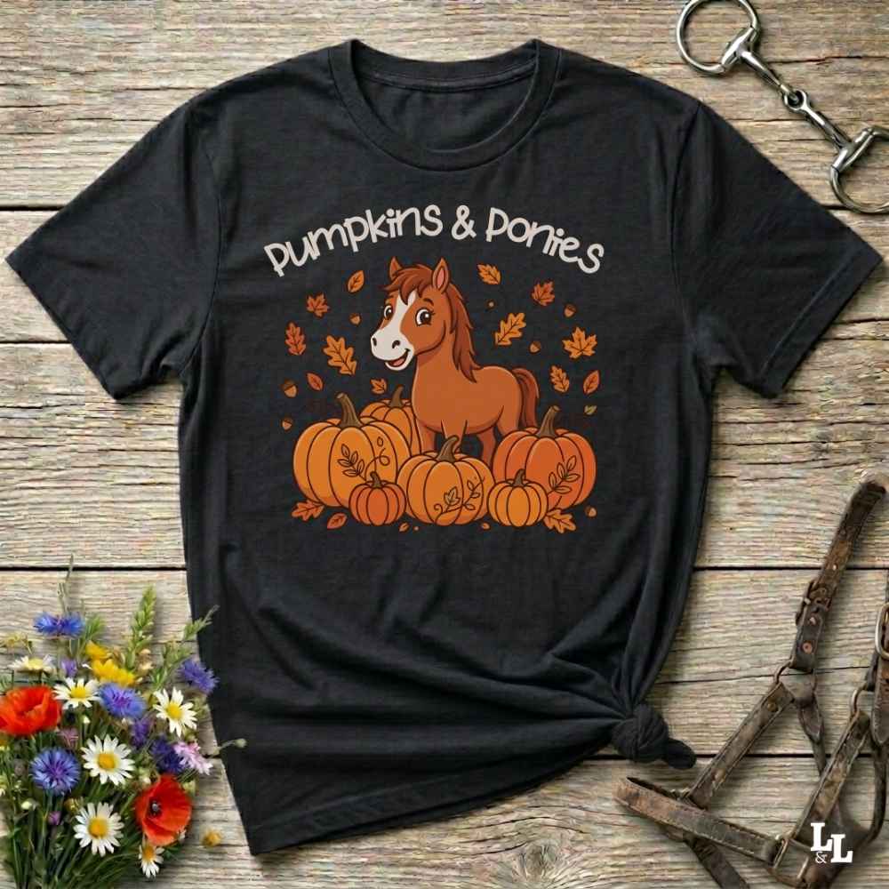 A black graphic t-shirt laid flat on a rustic wooden surface, featuring a cartoon brown pony sitting among orange pumpkins with the text 'Pumpkins & Ponies' above. The shirt is styled with a small bouquet of wildflowers on the left and horse tack props on the right.