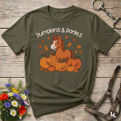 An olive green graphic t-shirt featuring a cartoon brown pony sitting among orange pumpkins with the text 'Pumpkins & Ponies' in white. The shirt is styled in a flat-lay on a rustic wooden background with a horse bit, leather halter, and a small bouquet of wildflowers.