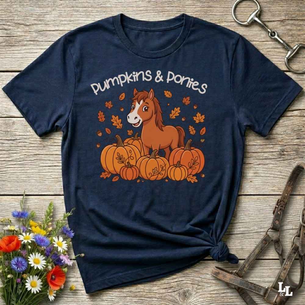 A rednavy blue short-sleeve graphic T-shirt laid flat on a rustic wooden surface, featuring a cartoon horse among pumpkins with the words 'Pumpkins & Ponies.' Decorative flowers and horse tack are placed around the shirt as styled props.