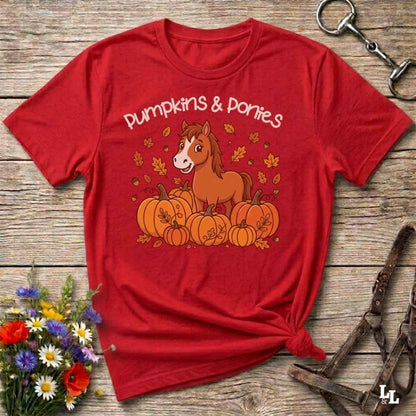 A red short-sleeve graphic T-shirt laid flat on a rustic wooden surface, featuring a cartoon horse among pumpkins with the words 'Pumpkins & Ponies.' Decorative flowers and horse tack are placed around the shirt as styled props.