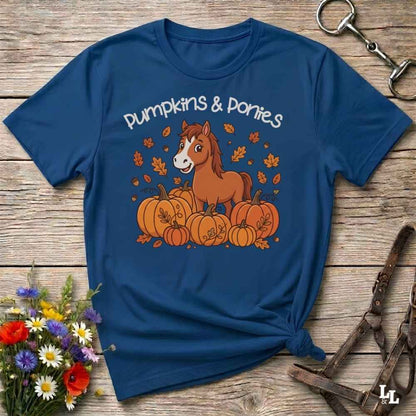 A navy blue graphic t-shirt featuring a cartoon pony and pumpkins with the text 'Pumpkins & Ponies' in white. The shirt is styled in a flat-lay on a rustic wooden background with a small bouquet of wildflowers and horse tack props.