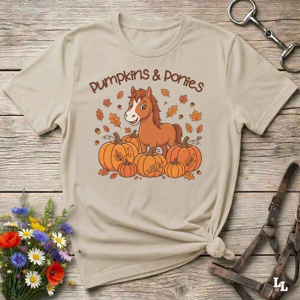 A beige graphic t-shirt featuring a cartoon pony among pumpkins with the text 'Pumpkins & Ponies' on the front, styled in a flat-lay on a rustic wooden background with decorative flowers and horse tack.