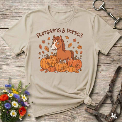 A beige graphic t-shirt featuring a cartoon pony among pumpkins with the text 'Pumpkins & Ponies' on the front, styled in a flat-lay on a rustic wooden background with decorative flowers and horse tack.