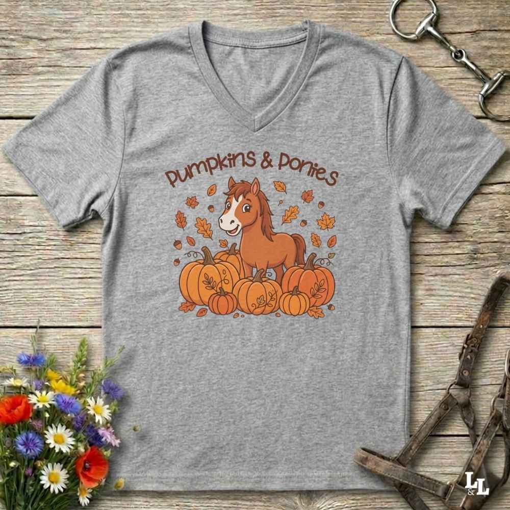 A heather gray V-neck t-shirt with a 'Pumpkins & Ponies' graphic, laid flat on a rustic wooden background with decorative autumn props.