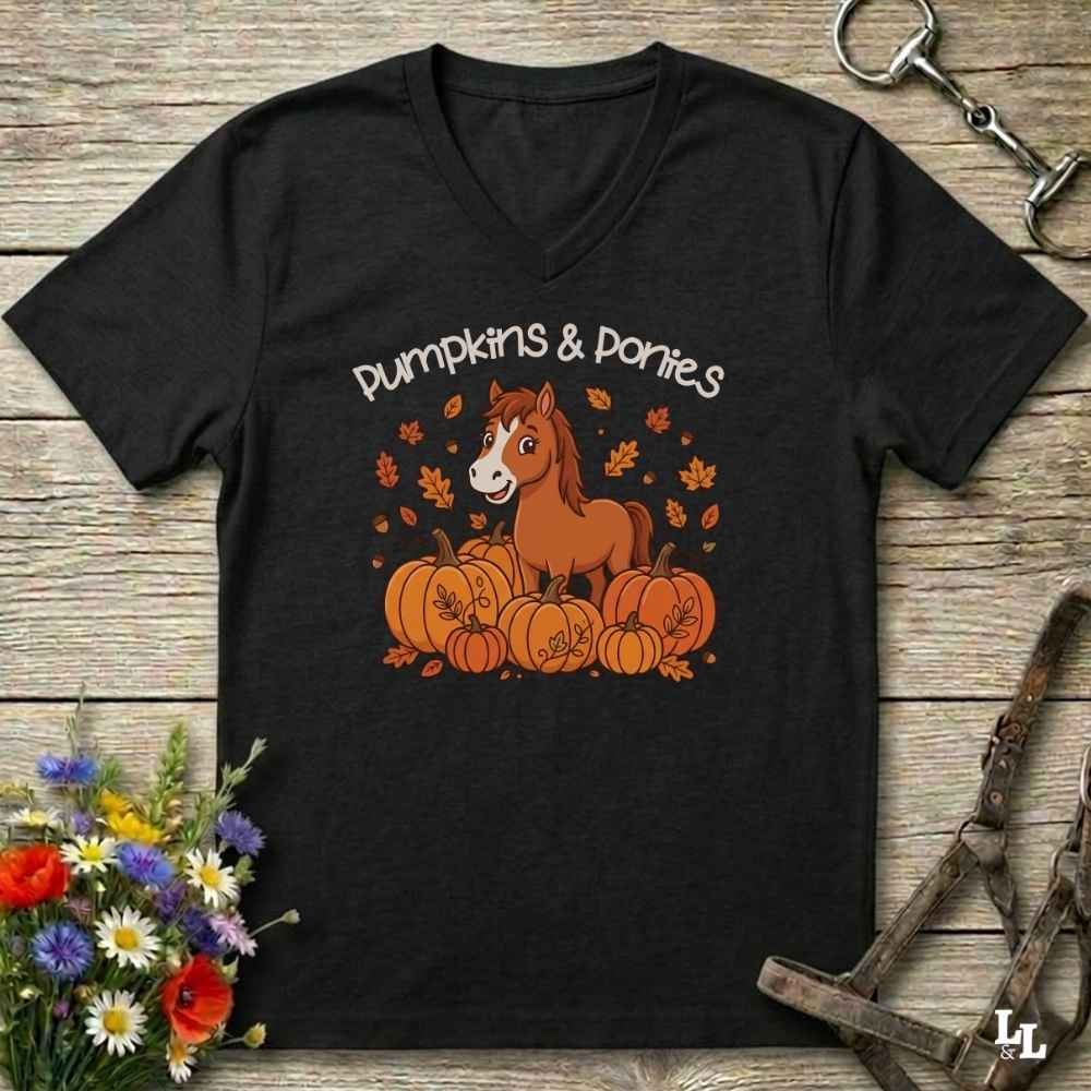 A black V-neck t-shirt with a 'Pumpkins & Ponies' graphic featuring a brown pony and pumpkins, laid flat on a rustic wooden background with decorative horse tack and flowers.