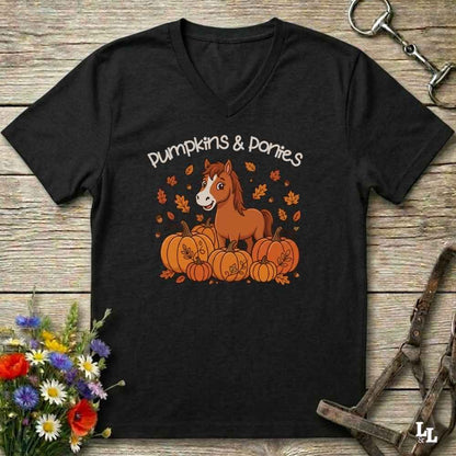 A black V-neck t-shirt with a 'Pumpkins & Ponies' graphic featuring a brown pony and pumpkins, laid flat on a rustic wooden background with decorative horse tack and flowers.