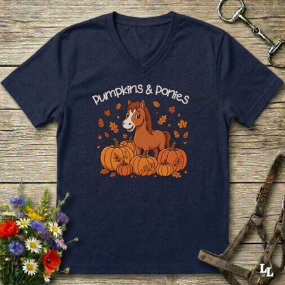 A navy blue V-neck t-shirt laid flat on a rustic wooden surface, featuring a graphic of a brown horse standing on orange pumpkins with the text 'Pumpkins & Ponies' above. The shirt is styled with equestrian tack in the background and a small bouquet of flowers in the lower left corner.