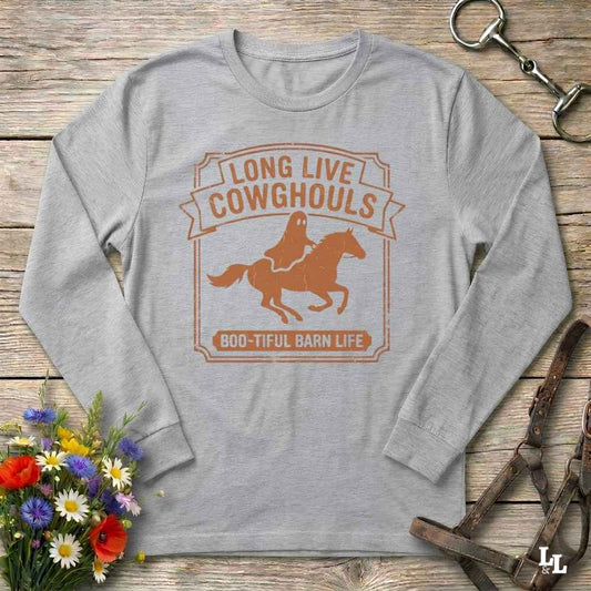 A light gray long-sleeve t-shirt with a brown graphic print of a ghost riding a horse and the text 'LONG LIVE COWGHOULS' and 'BOO-TIFUL BARN LIFE', displayed on a rustic wooden background with decorative props.