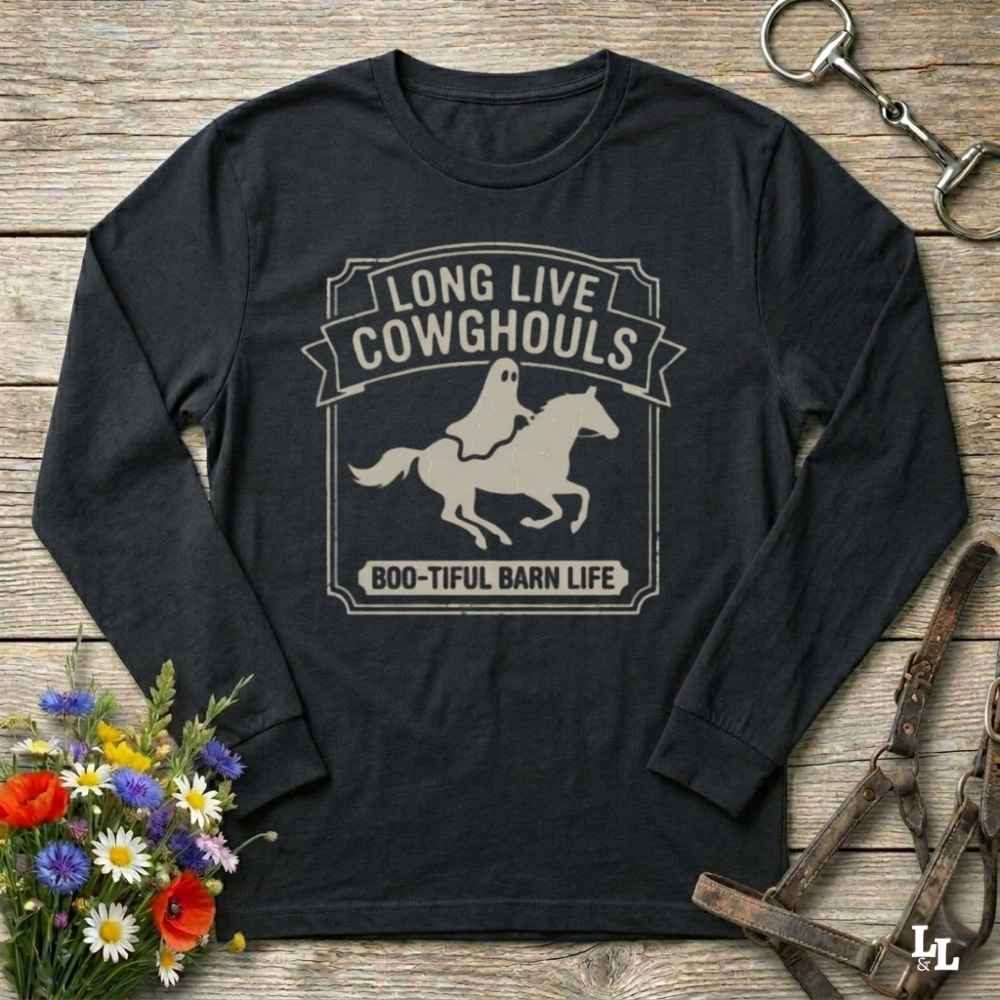 A black long-sleeve t-shirt with a distressed beige graphic that reads 'LONG LIVE COWGHOULS' and 'BOO-TIFUL BARN LIFE' surrounding a ghost riding a horse silhouette. The shirt is displayed flat on a rustic wooden background with Halloween-themed props including a bouquet of flowers, a horse bit, and a leather bridle.