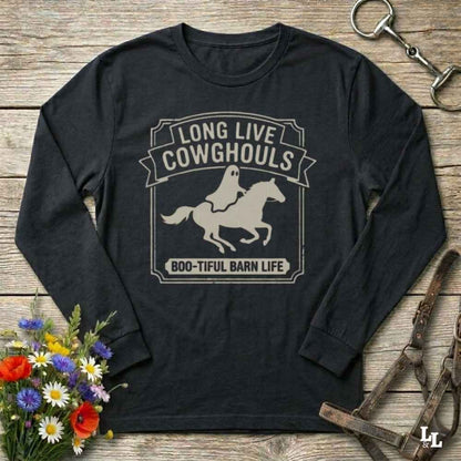 A black long-sleeve t-shirt with a distressed beige graphic that reads 'LONG LIVE COWGHOULS' and 'BOO-TIFUL BARN LIFE' surrounding a ghost riding a horse silhouette. The shirt is displayed flat on a rustic wooden background with Halloween-themed props including a bouquet of flowers, a horse bit, and a leather bridle.