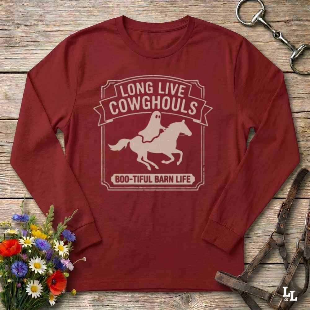 A long-sleeve crewneck t-shirt in maroon red, laid flat on a rustic wooden background. The shirt features a cream-colored graphic of a ghost riding a horse, with the text 'LONG LIVE COWGHOULS' and 'BOO-TIFUL BARN LIFE'.