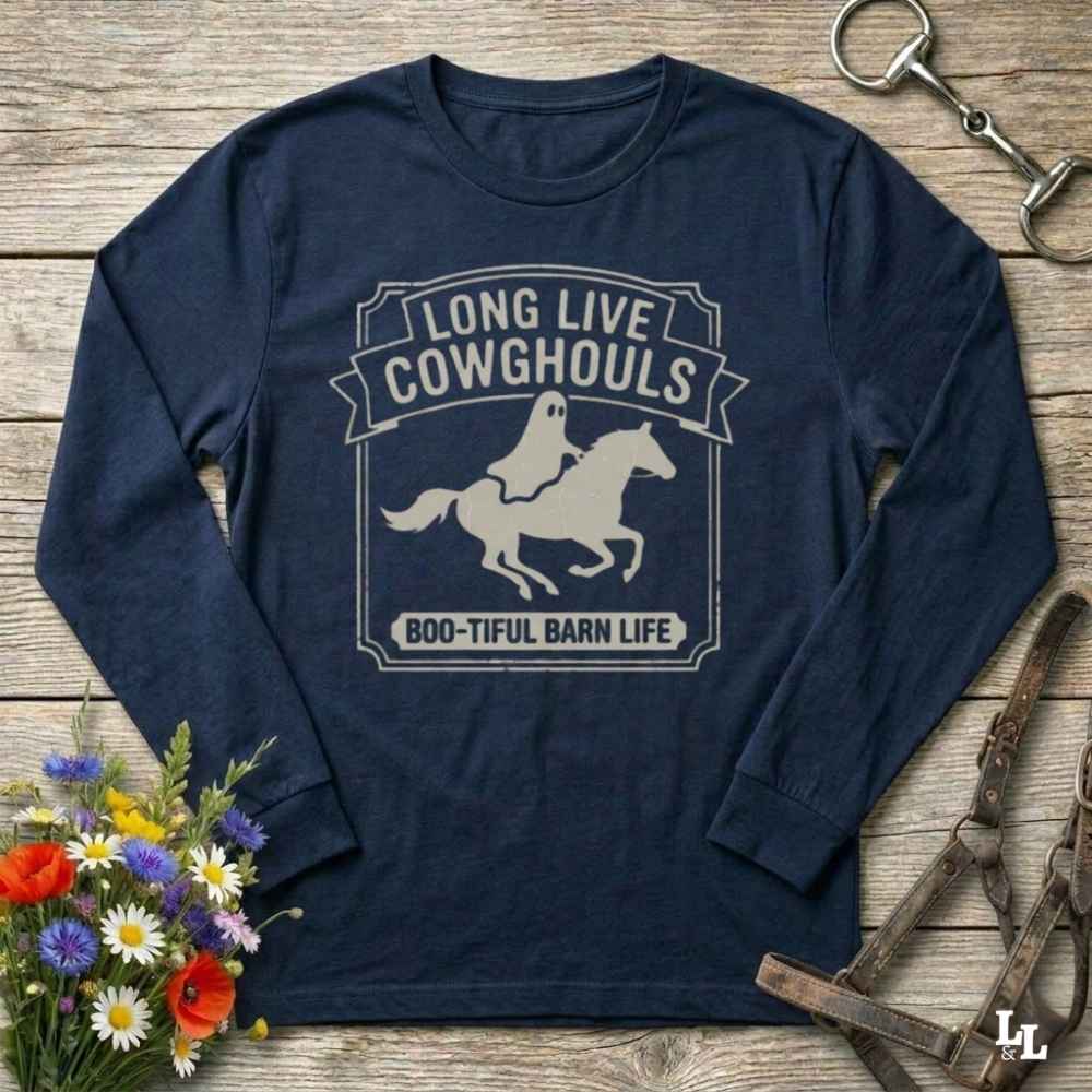 A navy blue long-sleeve t-shirt with a distressed white graphic reading 'LONG LIVE COWGHOULS' and 'BOO-TIFUL BARN LIFE' around a ghost riding a horse silhouette. The shirt is displayed in a flat-lay arrangement on a rustic wooden background, styled with a horse bit, leather bridle, and a small bouquet of wildflowers.