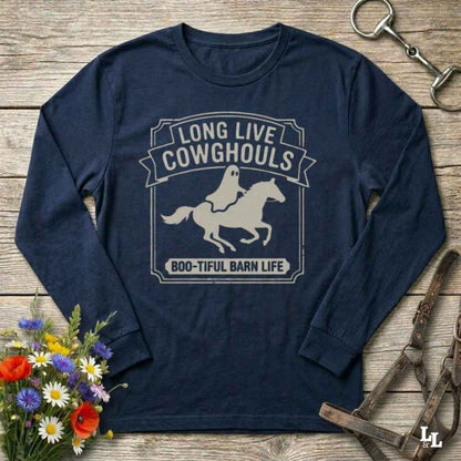 A navy blue long-sleeve t-shirt with a distressed white graphic reading 'LONG LIVE COWGHOULS' and 'BOO-TIFUL BARN LIFE' around a ghost riding a horse silhouette. The shirt is displayed in a flat-lay arrangement on a rustic wooden background, styled with a horse bit, leather bridle, and a small bouquet of wildflowers.