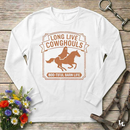 A white long-sleeve crewneck t-shirt with a brown graphic print of a ghost riding a horse and the text 'LONG LIVE COWGHOULS' and 'BOO-TIFUL BARN LIFE'. The shirt is displayed flat on a rustic wooden background with equestrian and floral props.