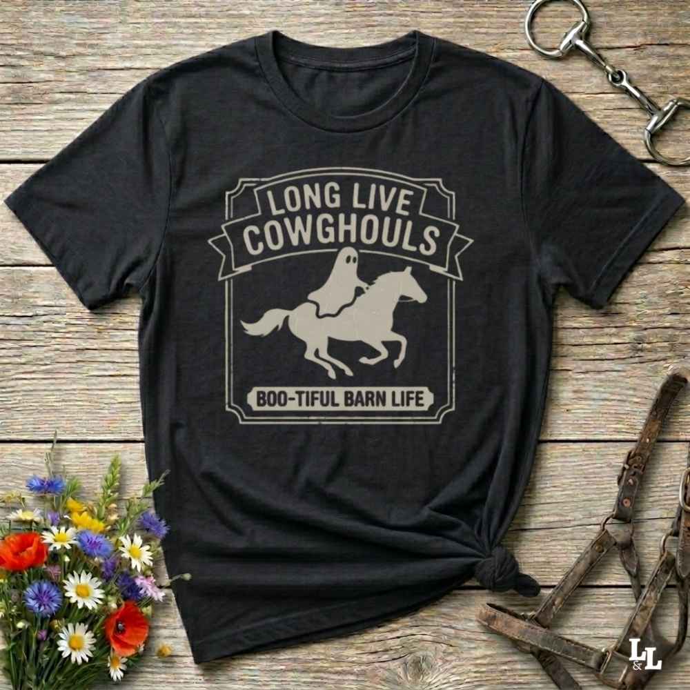 A black short-sleeve t-shirt with a distressed off-white graphic that reads 'LONG LIVE COWGHOULS' and 'BOO-TIFUL BARN LIFE' around a silhouette of a ghost riding a horse. The shirt is styled in a flat-lay on a rustic wooden background with equestrian accessories and a small bouquet of wildflowers.