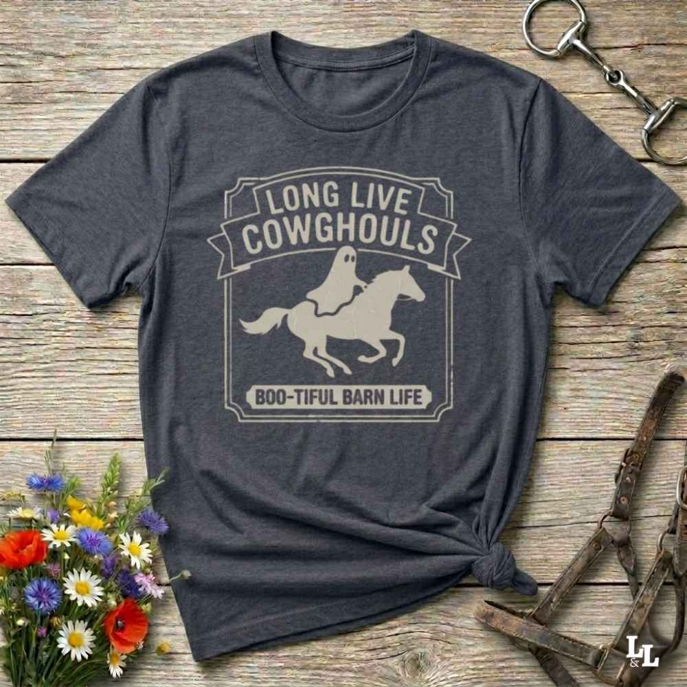 A dark heather short-sleeve t-shirt with a distressed off-white graphic that reads 'LONG LIVE COWGHOULS' and 'BOO-TIFUL BARN LIFE' around a silhouette of a ghost riding a horse. The shirt is styled in a flat-lay on a rustic wooden background with equestrian accessories and a small bouquet of wildflowers.