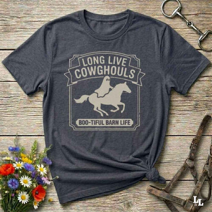A dark heather short-sleeve t-shirt with a distressed off-white graphic that reads 'LONG LIVE COWGHOULS' and 'BOO-TIFUL BARN LIFE' around a silhouette of a ghost riding a horse. The shirt is styled in a flat-lay on a rustic wooden background with equestrian accessories and a small bouquet of wildflowers.