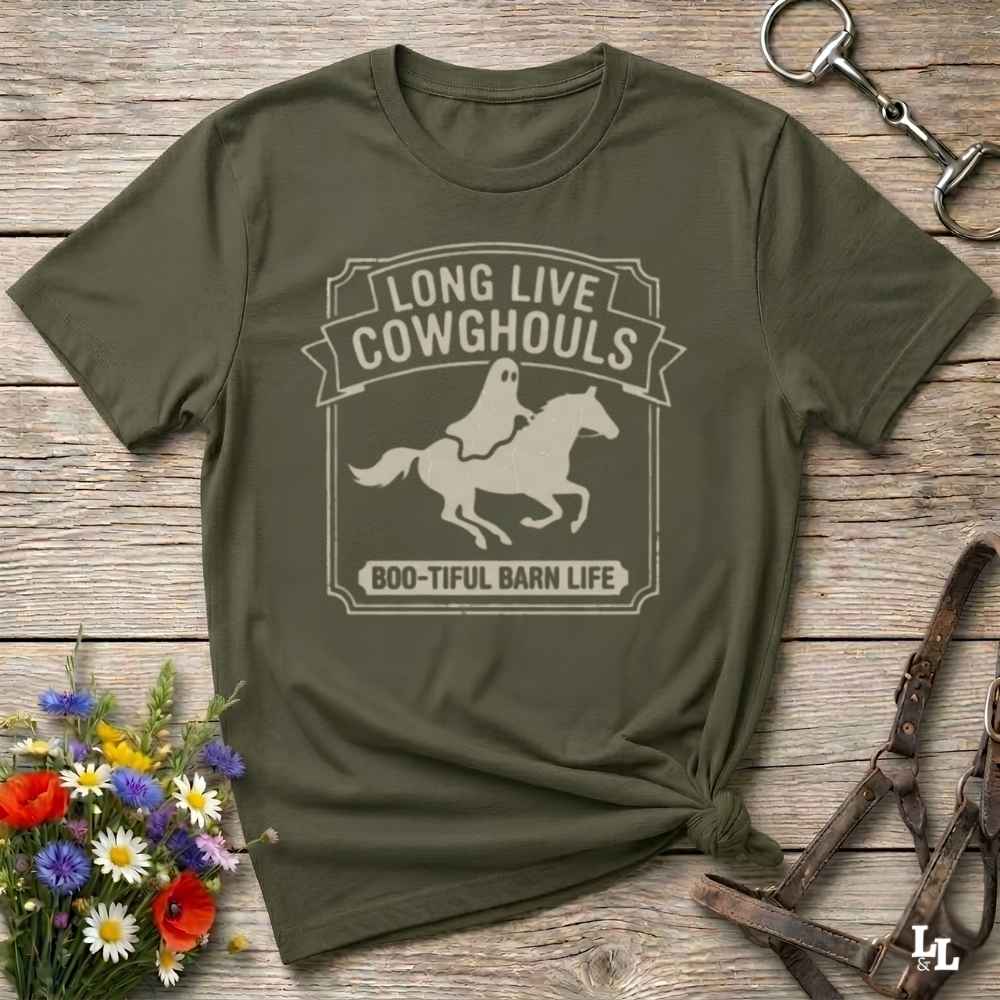 A green short-sleeve t-shirt with a distressed off-white graphic that reads 'LONG LIVE COWGHOULS' and 'BOO-TIFUL BARN LIFE' around a silhouette of a ghost riding a horse. The shirt is styled in a flat-lay on a rustic wooden background with equestrian accessories and a small bouquet of wildflowers.
