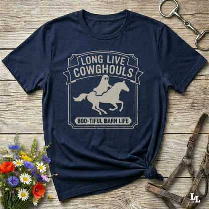 A navy blue short-sleeve t-shirt with a distressed off-white graphic that reads 'LONG LIVE COWGHOULS' and 'BOO-TIFUL BARN LIFE' around a silhouette of a ghost riding a horse. The shirt is styled in a flat-lay on a rustic wooden background with equestrian accessories and a small bouquet of wildflowers.