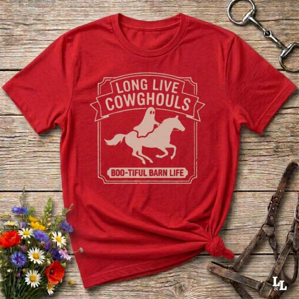 A red short-sleeve t-shirt with a distressed off-white graphic that reads 'LONG LIVE COWGHOULS' and 'BOO-TIFUL BARN LIFE' around a silhouette of a ghost riding a horse. The shirt is styled in a flat-lay on a rustic wooden background with equestrian accessories and a small bouquet of wildflowers.