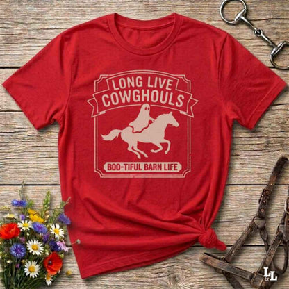 A red short-sleeve t-shirt with a distressed off-white graphic that reads 'LONG LIVE COWGHOULS' and 'BOO-TIFUL BARN LIFE' around a silhouette of a ghost riding a horse. The shirt is styled in a flat-lay on a rustic wooden background with equestrian accessories and a small bouquet of wildflowers.