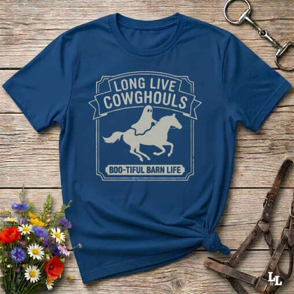 A blue short-sleeve t-shirt with a distressed off-white graphic that reads 'LONG LIVE COWGHOULS' and 'BOO-TIFUL BARN LIFE' around a silhouette of a ghost riding a horse. The shirt is styled in a flat-lay on a rustic wooden background with equestrian accessories and a small bouquet of wildflowers.