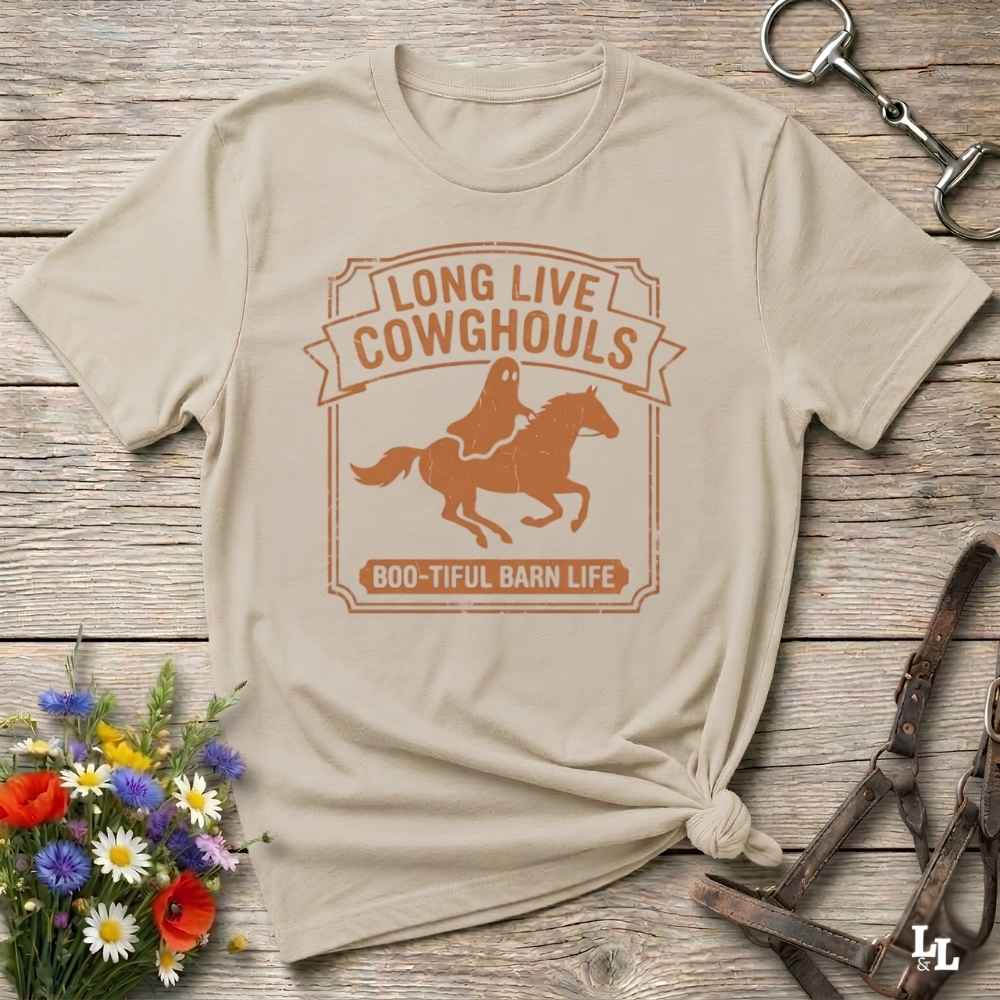 A beige short-sleeve t-shirt with a distressed off-white graphic that reads 'LONG LIVE COWGHOULS' and 'BOO-TIFUL BARN LIFE' around a silhouette of a ghost riding a horse. The shirt is styled in a flat-lay on a rustic wooden background with equestrian accessories and a small bouquet of wildflowers.
