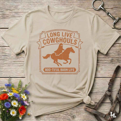 A beige short-sleeve t-shirt with a distressed off-white graphic that reads 'LONG LIVE COWGHOULS' and 'BOO-TIFUL BARN LIFE' around a silhouette of a ghost riding a horse. The shirt is styled in a flat-lay on a rustic wooden background with equestrian accessories and a small bouquet of wildflowers.