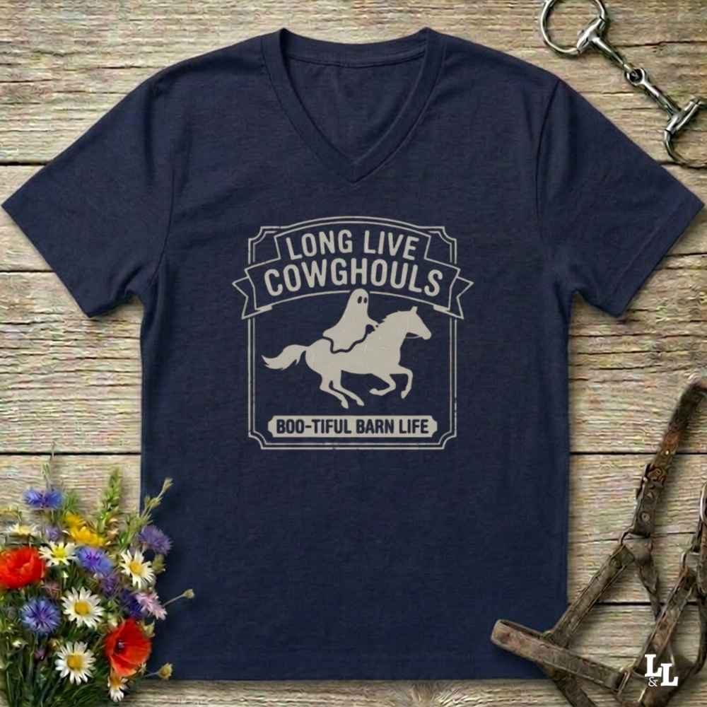 A navy blue V-neck t-shirt with a white graphic print reading 'LONG LIVE COWHOOLS' and 'BOO-TIFUL BARN LIFE' around a ghost riding a horse. The shirt is laid flat on a rustic wooden background with equestrian and floral props.