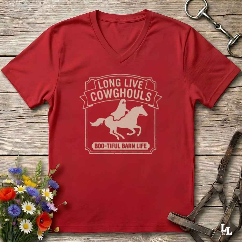 A red V-neck t-shirt laid flat on a rustic wooden surface, featuring a cream-colored graphic of a ghost riding a horse with the text 'LONG LIVE COWHOULDS' and 'BOO-TIFUL BARN LIFE'. The shirt is styled with equestrian tack in the background and a small bouquet of wildflowers in the lower-left corner.