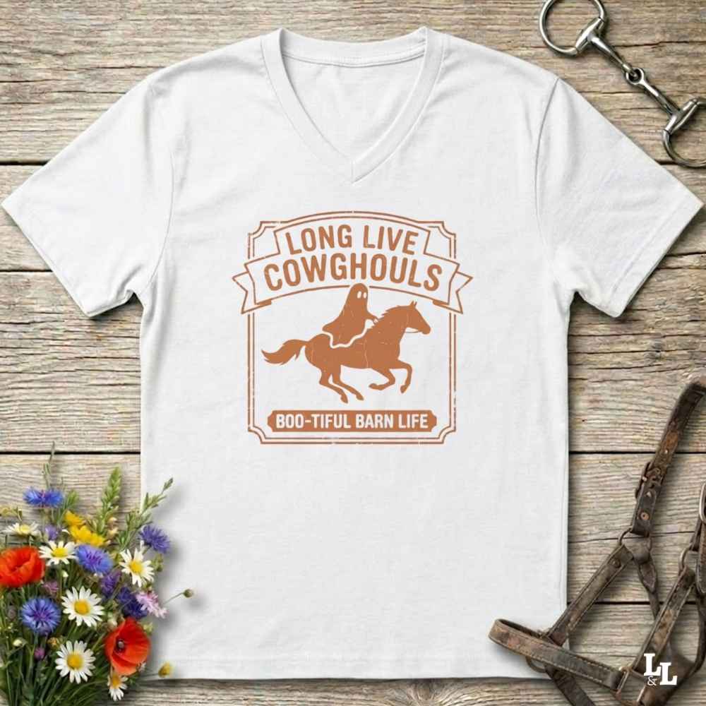 A white V-neck t-shirt laid flat on a rustic wooden surface, featuring a brown graphic of a cowboy riding a horse with the text 'LONG LIVE COWHOULS' and '800-TIFUL BARN LIFE'. The shirt is styled with equestrian and floral props.