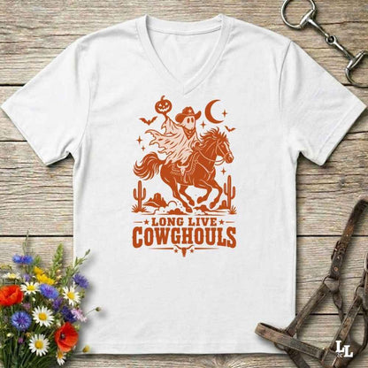 A white V-neck t-shirt with a burnt orange graphic of a skeleton cowboy riding a horse through a desert scene with the text 'LONG LIVE COWGHOULS'. The shirt is displayed flat on a rustic wooden background with equestrian and floral props.
