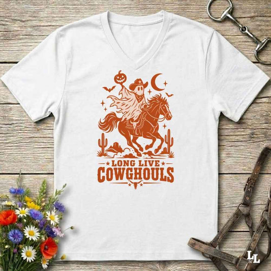 A white V-neck t-shirt with a burnt orange graphic of a skeleton cowboy riding a horse through a desert scene with the text 'LONG LIVE COWGHOULS'. The shirt is displayed flat on a rustic wooden background with equestrian and floral props.