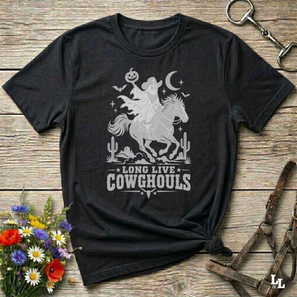 A black short-sleeve graphic T-shirt laid flat on a rustic wooden surface, with a white western-themed print showing a cowboy on a horse, cacti, stars, and the text 'LONG LIVE COWGHOULS.' Decorative flowers and leather tack are placed around the shirt as props.