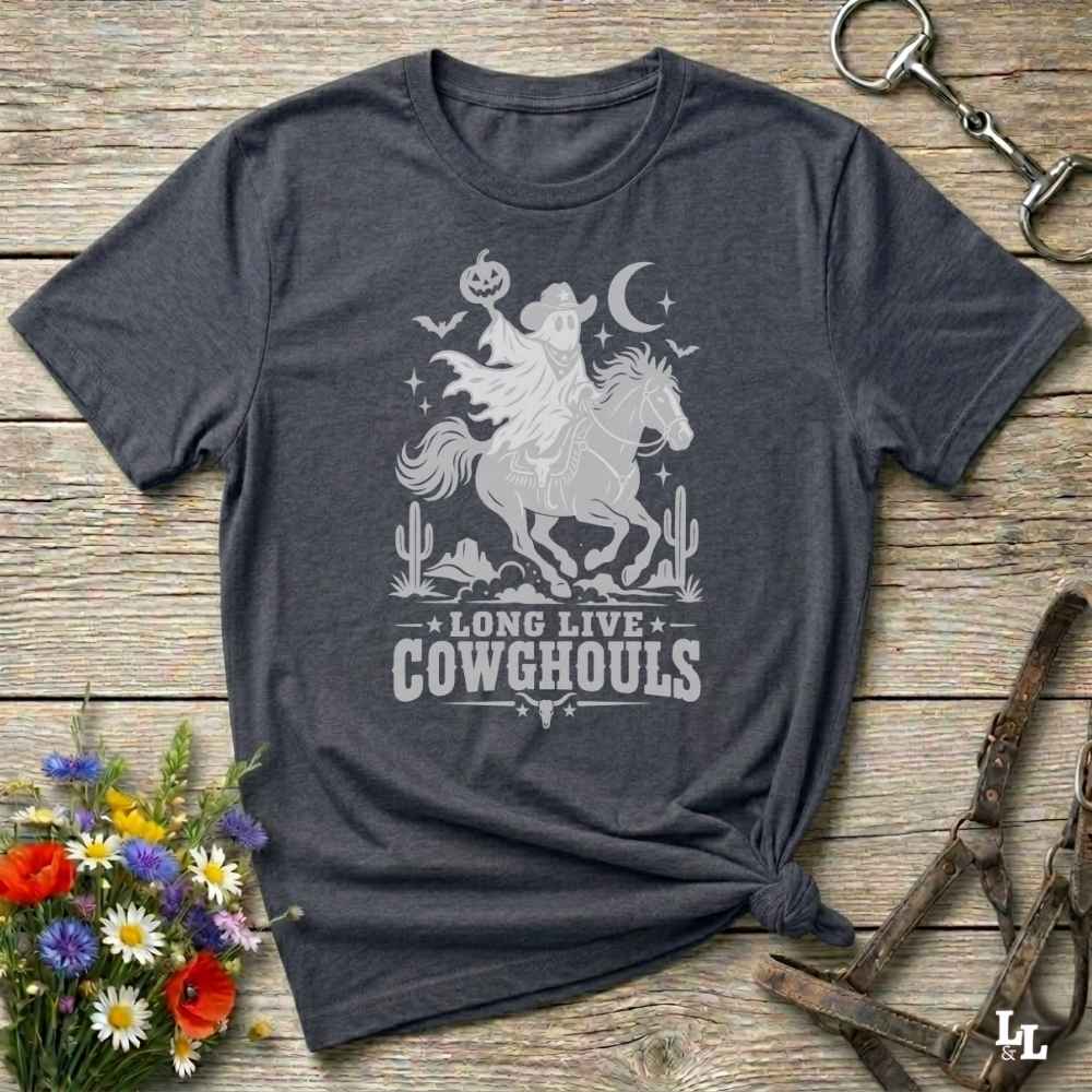 A grey short-sleeve graphic T-shirt laid flat on a rustic wooden surface, with a white western-themed print showing a cowboy on a horse, cacti, stars, and the text 'LONG LIVE COWGHOULS.' Decorative flowers and leather tack are placed around the shirt as props.