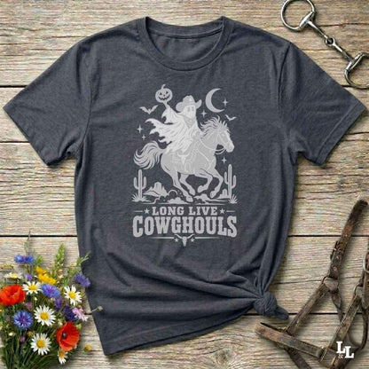A grey short-sleeve graphic T-shirt laid flat on a rustic wooden surface, with a white western-themed print showing a cowboy on a horse, cacti, stars, and the text 'LONG LIVE COWGHOULS.' Decorative flowers and leather tack are placed around the shirt as props.