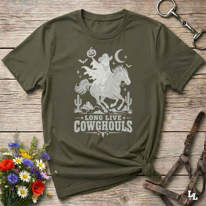 A green  short-sleeve graphic T-shirt laid flat on a rustic wooden surface, with a white western-themed print showing a cowboy on a horse, cacti, stars, and the text 'LONG LIVE COWGHOULS.' Decorative flowers and leather tack are placed around the shirt as props.