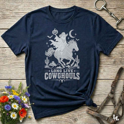 A navy blue graphic t-shirt featuring a white western-themed design with a cowboy on a horse, cacti, a crescent moon, and the text 'LONG LIVE COWGHOULS'. The shirt is styled in a flat-lay on a rustic wooden background with decorative flowers and leather tack.