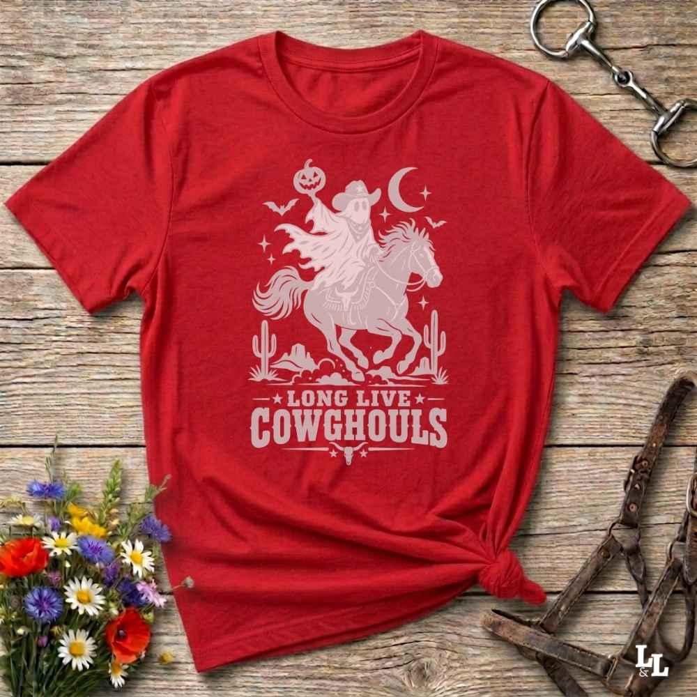 A red short-sleeve graphic T-shirt laid flat on a rustic wooden surface, with a white cowboy silhouette print reading 'LONG LIVE COWGHOULS' and decorative western motifs. The shirt is styled with flowers, a horse bit, and a leather bridle around it.