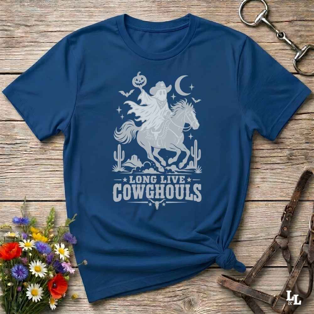 A navy blue short-sleeve t-shirt with a white graphic of a cowboy riding a horse, surrounded by a moon, stars, cacti, and the text 'LONG LIVE COWGHOULS'. The shirt is styled in a flat-lay on a rustic wooden background with decorative horse tack and a small bouquet of wildflowers.