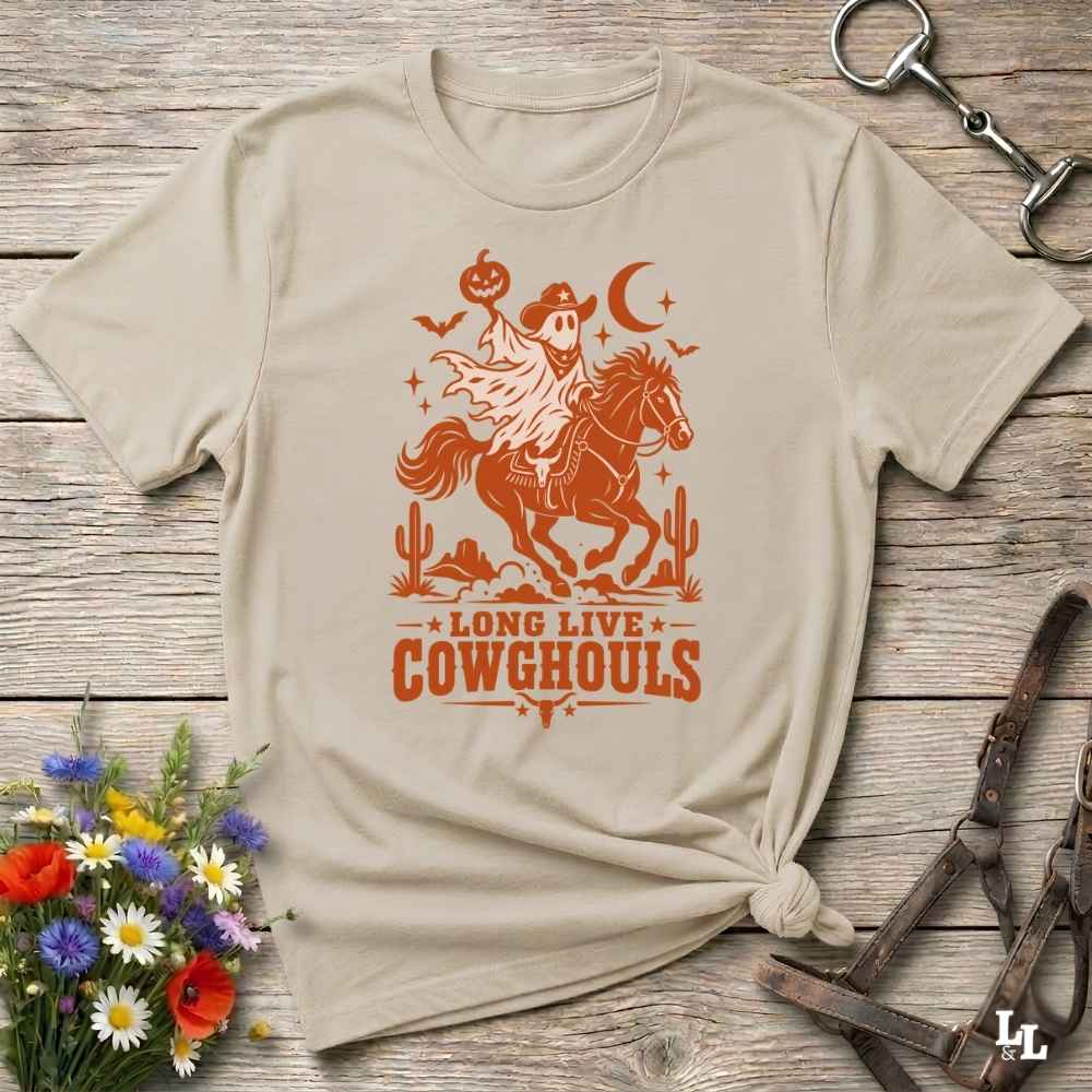A beige short-sleeve graphic T-shirt laid flat on a rustic wooden surface, with a white western-themed print showing a cowboy on a horse, cacti, stars, and the text 'LONG LIVE COWGHOULS.' Decorative flowers and leather tack are placed around the shirt as props.