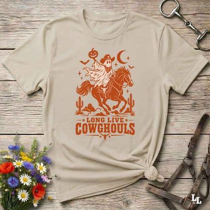 A beige short-sleeve graphic T-shirt laid flat on a rustic wooden surface, with a white western-themed print showing a cowboy on a horse, cacti, stars, and the text 'LONG LIVE COWGHOULS.' Decorative flowers and leather tack are placed around the shirt as props.