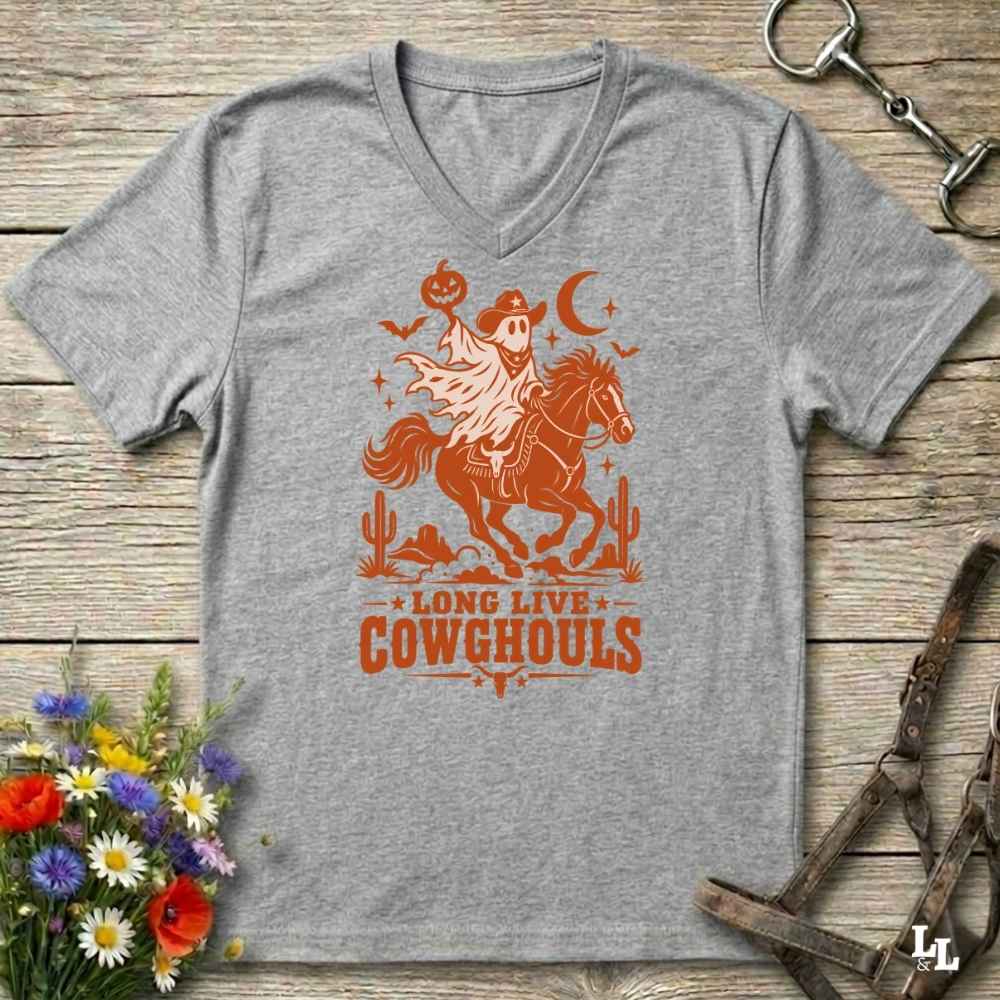 A grey V-neck t-shirt with a burnt orange graphic of a skeleton cowboy riding a horse through a desert scene with the text 'LONG LIVE COWGHOULS'. The shirt is displayed flat on a rustic wooden background with equestrian and floral props.